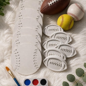 May include: A collection of sports-themed craft supplies. Includes white baseball-shaped coasters, football and baseball models, a yellow softball, a watercolor paint palette, and a paintbrush. The items are arranged on a fluffy white surface.