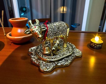 May include: A gold-colored cow figurine with intricate detailing, a red ribbon, and a small bell. The cow stands on an ornate base, next to a small candle and a terracotta pot. The scene is set on a wooden surface.