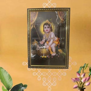 May include: A framed religious print of a young boy, possibly Krishna, holding a pot of milk. The print is set against a dark background with a golden halo around the boy's head. The frame is black with gold accents.