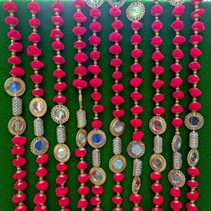 May include: Rows of red pom-poms and decorative mirrored and gold-toned ornaments hang against a green background. The ornaments vary in shape and size, creating a festive and eye-catching display. The design is symmetrical and visually appealing.