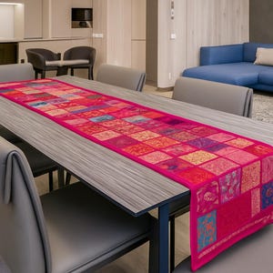May include: A vibrant, patchwork-style table runner in shades of pink, red, and orange. The rectangular runner is placed on a gray wooden table, with matching chairs. The runner's design features a variety of patterned squares.