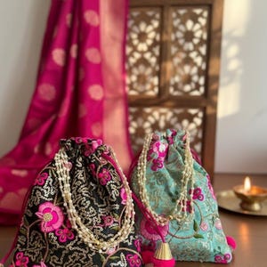 May include: Two drawstring bags with floral embroidery. One bag is black with pink flowers and gold sequins, the other is teal with pink flowers. Both have beaded accents and pink tassels. A pink sari is in the background.
