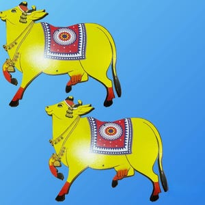May include: Two yellow wooden cow figurines with red and blue patterned saddle blankets. The cows are standing on a blue background.