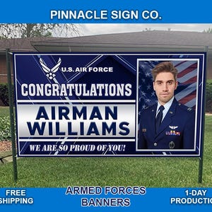 May include: A blue and white banner with the text "U.S. AIR FORCE" and "CONGRATULATIONS AIRMAN WILLIAMS WE ARE SO PROUD OF YOU!" A photo of a man in a military uniform is in the center of the banner. The banner is hanging from a metal pole.