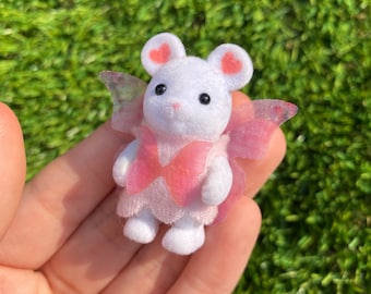 May include: A white mouse figurine with pink heart-shaped ears and a pink bow. The mouse has black eyes and is wearing a pink dress with translucent wings. The figurine is a toy.