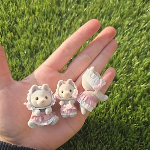 Puede incluir: Tres figuras de osos en miniatura con vestidos y gorros rosas y blancos. Los osos tienen pelaje gris y están posados sobre una mano sobre un fondo de hierba verde. Cada oso tiene una pose diferente.