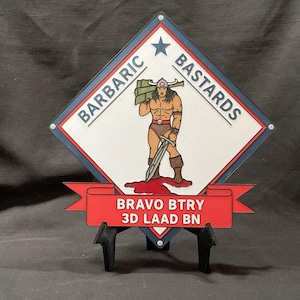 May include: A wooden sign featuring a muscular warrior with a sword, the words "BARBARIC BASTARDS" and "BRAVO BTRY 3D LAAD BN". The sign is in a diamond shape with a red banner and is mounted on a black stand.
