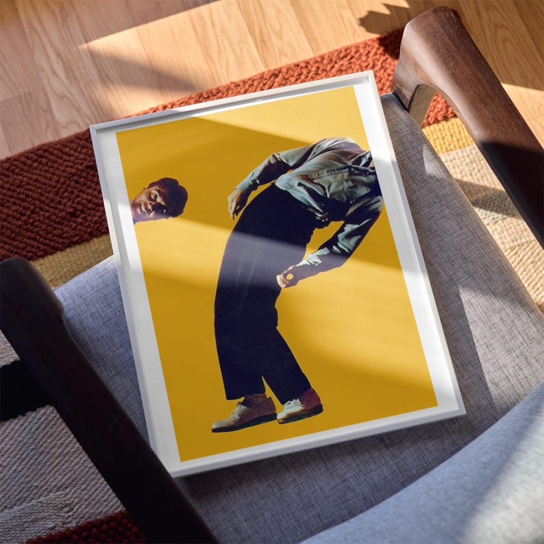 May include: A framed art print featuring a person in a light-colored long-sleeve shirt and dark trousers bending forward against a solid yellow background. A partial view of a face is visible on the left side of the frame, looking toward the center.