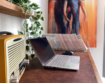 May include: A silver laptop sits on a dark brown leather desk mat next to a yellow vintage-style radio with a cream-colored speaker grille and two silver knobs. A trailing green plant hangs from a wooden shelf above the desk area.
