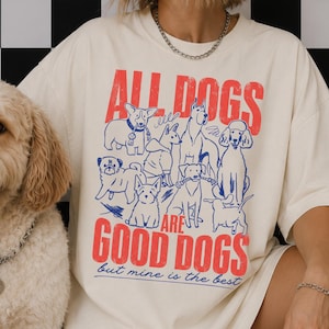 May include: An off-white t-shirt featuring a graphic print of various dog breeds in blue outlines. The text in red and blue reads: ALL DOGS ARE GOOD DOGS but mine is the best. The design includes a variety of canine illustrations.