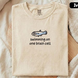May include: Ivory-colored t-shirt with a cartoon fish illustration and the text "swimming on one brain cell." The fish is blue and black. The shirt is folded and placed on a wooden surface.