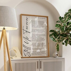 May include: A framed print with the words "Still Us Forever" in elegant script. The print features a poem with the names "Kaylee & Lennon" at the top. The frame is a light brown colour, and the print is displayed on a white cabinet.