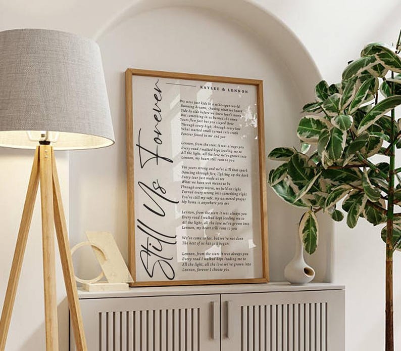 May include: A framed print with the words "Still Us Forever" in elegant script. The print features a poem with the names "Kaylee & Lennon" at the top. The frame is a light brown colour, and the print is displayed on a white cabinet.