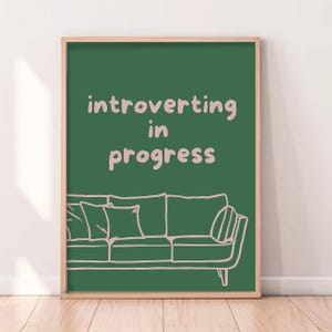 May include: A framed art print with a green background and the words "introverting in progress" in light pink. A line drawing of a sofa is at the bottom of the print. The frame is a light brown color.