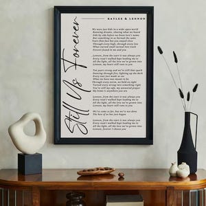May include: A framed print with the words "Still Us Forever" in elegant script. The print features a poem with the names "Kaylee & Lennon" at the top. The artwork is displayed on a wooden cabinet with decorative items.