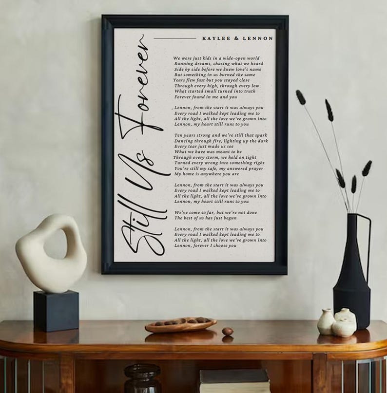 May include: A framed print with the words "Still Us Forever" in elegant script. The print features a poem with the names "Kaylee & Lennon" at the top. The artwork is displayed on a wooden cabinet with decorative items.