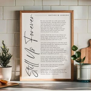 May include: A framed print with the words "Still Us Forever" in elegant script. The print features a poem with the names "Kaylee & Lennon" at the top. The frame is a light brown colour, and the background is white.