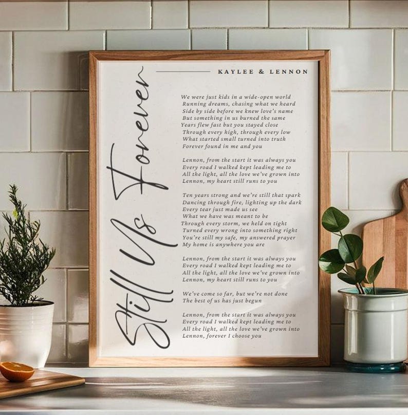May include: A framed print with the words "Still Us Forever" in elegant script. The print features a poem with the names "Kaylee & Lennon" at the top. The frame is a light brown colour, and the background is white.