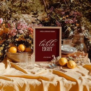 May include: A burgundy sign with the words "table EIGHT" in a wooden frame, part of a wedding table setting. The scene includes lemons, glasses, and cutlery on a yellow tablecloth. The sign also reads "Amelia & Benjamin Wedding" and "22nd of June 2025".
