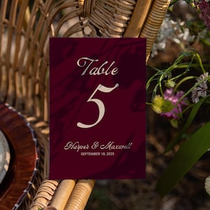 May include: A burgundy table number card with the text "Table 5" in an elegant script. The card also includes the names "Harper & Maxwell" and the date "September 18, 2025". The card is set against a blurred background of flowers and a wicker chair.