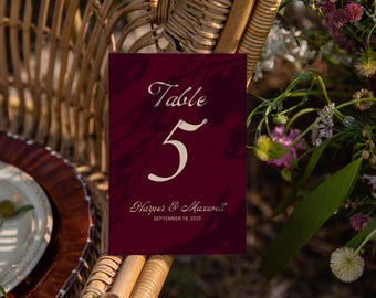 May include: A burgundy table number card with the text "Table 5" in an elegant script. The card also includes the names "Harper & Maxwell" and the date "September 18, 2025". The card is set against a blurred background of flowers and a wicker chair.