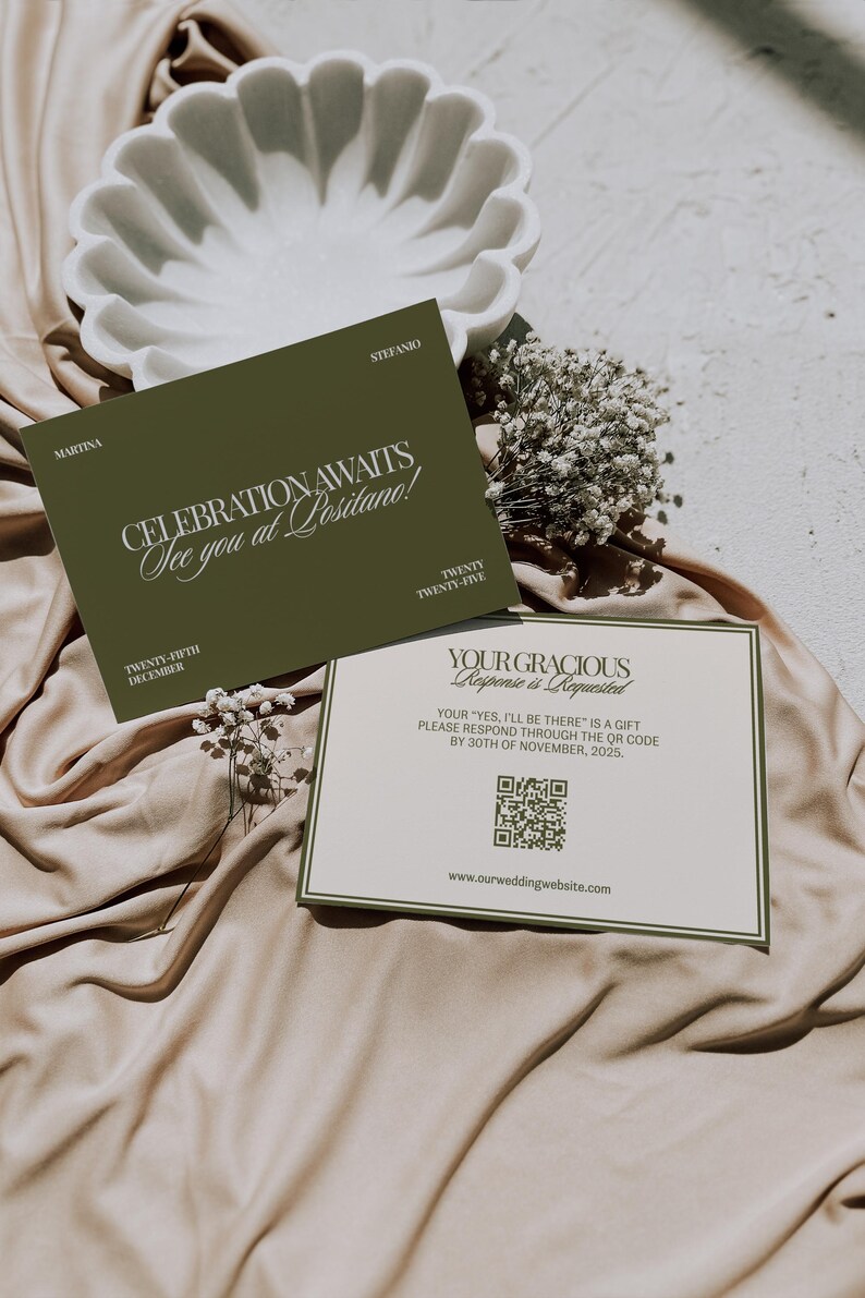 Two olive green and cream wedding stationery cards rest on satin fabric near a decorative white bowl and white flowers.