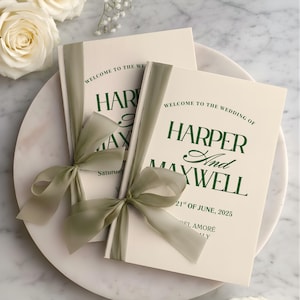 Pode incluir: Dois programas de casamento com laços de fita verde sálvia. Os programas creme apresentam o texto verde escuro: "WELCOME TO THE WEDDING OF HARPER And MAXWELL" e a data "21st OF JUNE, 2025". Estão sobre um prato de mármore.
