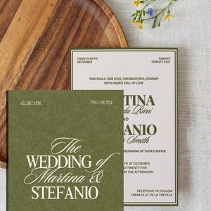 An olive green and cream wedding invitation set is displayed next to a wooden plate and small yellow and blue flowers.