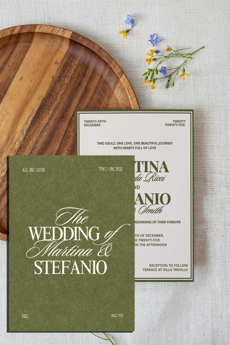 An olive green and cream wedding invitation set is displayed next to a wooden plate and small yellow and blue flowers.