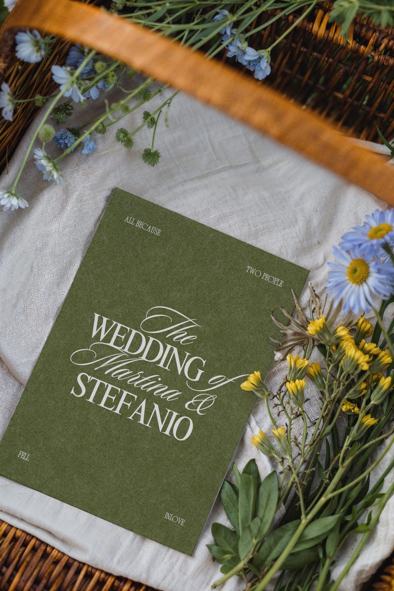 An olive green wedding program card with white text rests in a basket with blue and yellow flowers on cloth.