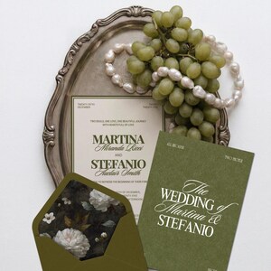 An olive green and cream wedding invitation suite rests on a silver tray with green grapes and a string of pearls.