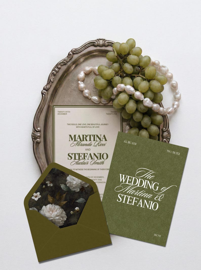 An olive green and cream wedding invitation suite rests on a silver tray with green grapes and a string of pearls.