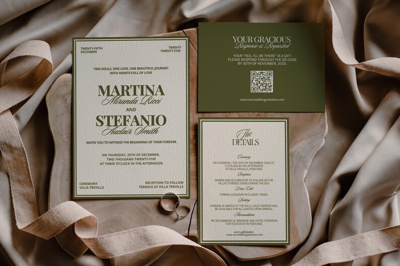 Olive green and cream wedding stationery cards with two silver rings rest on folded draped fabric.