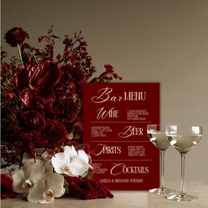 May include: A dark red bar menu sign with white text listing wine, beer, spirits, and cocktails for the Amelia & Benjamin wedding, accompanied by two clear glass coupes and a large arrangement of deep red roses and white orchids.