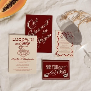 May include: A collection of wedding invitation cards with a Las Vegas theme. The cards are in shades of cream and burgundy, with text that reads "Lucky in Love" and "See You in Las Vegas". A slice of papaya and a glass are also in the image.