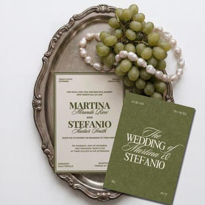 An olive green and cream wedding invitation suite sits on an ornate silver tray next to a bunch of green grapes and pearls.