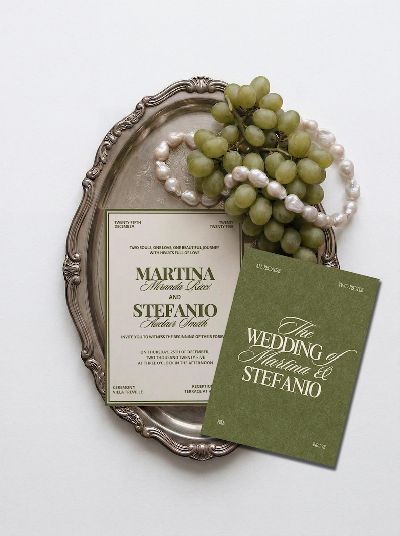 An olive green and cream wedding invitation suite sits on an ornate silver tray next to a bunch of green grapes and pearls.