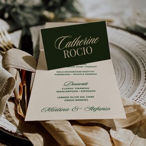 Könnte beinhalten: Eine Hochzeitsmenükarte mit den Namen "Catherine Rocio" in eleganter Schrift. Das Menü listet Gerichte wie Wildpilzrisotto und klassisches Tiramisu auf. Die Karte liegt auf einem Tisch mit einer beigen Serviette und einem dekorativen Teller.
