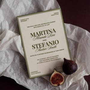 A cream and olive green wedding invitation card rests on white tissue paper with a whole fig and a sliced fig.