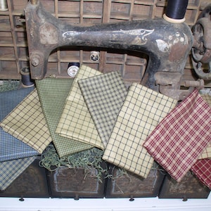 May include: Assortment of six fabric squares in various plaid patterns. The fabrics are in shades of green, blue, red, and beige. The fabrics are arranged in a fan shape and are displayed on top of a wooden box.
