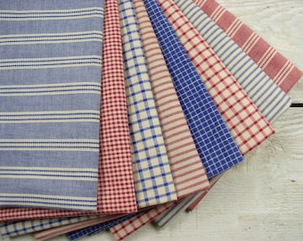 May include: A collection of folded fabric cloths in various patterns and colors. The cloths feature stripes, checks, and other designs in shades of blue, red, and cream. The fabrics are arranged in a fan shape, showcasing the variety of patterns.