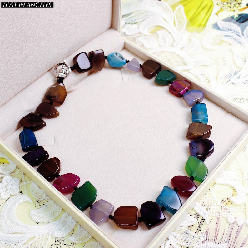 May include: A multi-coloured gemstone necklace with a silver clasp, displayed in a white box. The necklace features polished, irregularly shaped stones in shades of brown, blue, green, purple, and red. The text "LOST IN ANGELES" is in the top left corner.
