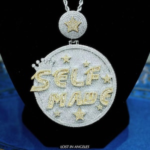 May include: A silver pendant necklace featuring the words "SELF MADE" in gold-coloured, gem-studded letters. The pendant has a circular design with star accents and a crown. A smaller, star-shaped pendant is at the top. The text "LOST IN ANGELES" is at the bottom.