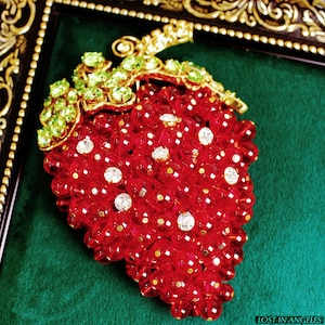 May include: A red beaded strawberry brooch with clear and gold accents. The brooch features a gold stem and green gemstone leaves. The strawberry is set against a green velvet background and framed in gold.
