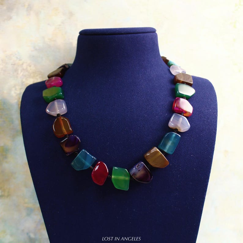 May include: A vibrant gemstone necklace on a blue display. The necklace is made of irregularly shaped stones in red, green, blue, brown, pink, and purple. The stones are strung together with a black cord.