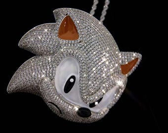 May include: A pendant necklace featuring a sparkling, diamond-encrusted Sonic the Hedgehog design. The pendant has brown accents on the ears and a white face with black eyes and nose. The necklace hangs on a white chain against a black background.