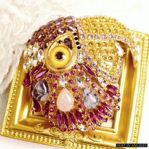May include: A gold-coloured owl brooch, adorned with an array of gemstones. The brooch showcases a prominent yellow eye, encircled by purple, pink, and clear crystals. The owl is set within an ornate, gold-toned frame. The text "LOST IN ANGELS" is visible.