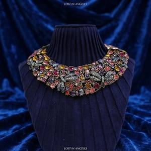 May include: A statement collar necklace featuring a dense arrangement of multicoloured round crystals in shades of yellow, pink, purple, and blue, accented with metallic dragonfly motifs. The text LOST IN ANGELES appears at the top and bottom.