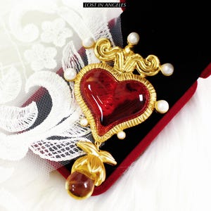 May include: A gold-toned heart-shaped pendant with a red center, accented with faux pearls. The pendant is set against a black velvet background with the text "LOST IN ANGELES" at the top. A gold-toned floral element hangs below the heart.