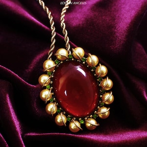 May include: A gold-toned necklace with a large, oval, red gemstone pendant. The pendant is surrounded by gold-colored beads and small green stones. The necklace chain is a twisted gold rope. The text "LOS N ANGELES" is visible in the upper right corner.
